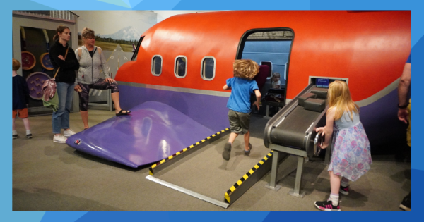 A child runs up a ramp into the open door of a children's museum exhibit designed to look like an airplane, while a girl cranks a conveyor belt loaded with luggage. 