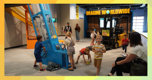 Interior of a children's museum where children gather around and place boxes into a conveyance system with a modified forklift in the background.