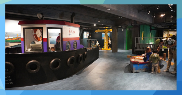 An interior scene in a children's museum, with a pretend tug boat, crane and row boat.